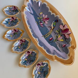 May include: A set of vintage serving dishes. The set includes a large oval serving dish and six smaller leaf-shaped dishes. Each dish has a gold rim and features a floral and bird design in shades of pink, blue, and yellow.