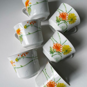 May include: Six white teacups with floral designs. The cups feature a white background with green stems and orange and yellow flowers.