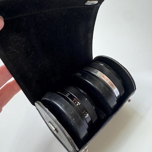 May include: Black leather camera lens case with two black lenses inside. The lenses have white and red markings. The case is open, revealing the lenses. The case is rectangular with a snap closure.