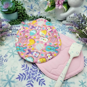 Puede incluir: Dos posavasos acolchados de color rosa con un patrón de cuadros rosa y blanco. Un posavasos tiene un estampado colorido con temática de Pascua con conejos, pollitos y huevos. Una espátula de silicona blanca con motas rosas y azules está encima de los posavasos.