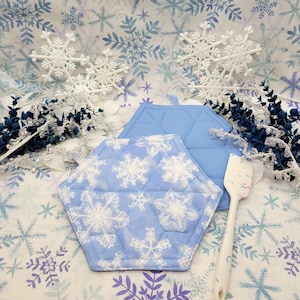 Puede incluir: Dos agarraderas de olla acolchadas azules con un patrón de copo de nieve. Las agarraderas de olla son hexagonales y tienen un diseño de copo de nieve blanco sobre un fondo azul claro. Las agarraderas de olla están sobre un fondo blanco con un patrón de copo de nieve.