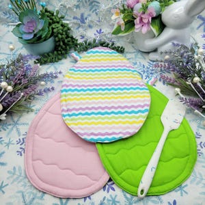 Puede incluir: Tres posavasos acolchados de colores con forma de huevo. Los posavasos son rosa, verde y rayados de amarillo, azul y morado. Una espátula de silicona blanca está encima del posavasos verde.