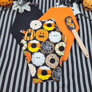 May include: Two black and orange Halloween-themed pot holders, one shaped like a coffin and the other a traditional hexagon. The coffin pot holder features a pattern of donuts with Halloween-themed decorations, such as pumpkins, spiderwebs, and candy corn. The hexagon pot holder is plain black. A wooden spatula with a black handle and a Halloween-themed design is also included.