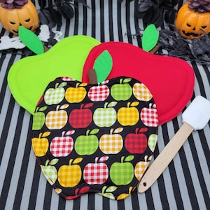 May include: Three apple-shaped pot holders in green, red, and a black fabric with apple patterns. A white spatula with a wooden handle rests on the striped background. Decorative pumpkins are in the background.