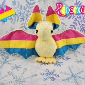 May include: A plush bat toy with pink, yellow, and blue wings, representing the pansexual pride flag. The bat is cream colored and has a cute, cartoonish face.