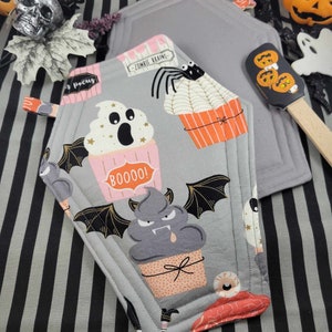 May include: A gray and white Halloween-themed pot holder shaped like a coffin with a variety of cupcakes decorated with ghosts, bats, and spiders. The cupcakes have pink and orange frosting and are decorated with sprinkles and stars. The text "Boo!" and "Zombie Brains" are printed on the pot holder.