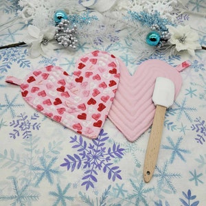 Könnte beinhalten: Zwei rosafarbene herzförmige Topflappen mit einem Holzlöffel auf einem weißen Hintergrund mit Schneeflockenmuster. Ein Topflappen hat ein rosa-rotes Muster mit dem Wort "love", das darauf wiederholt wird. Der andere Topflappen ist einfarbig rosa.