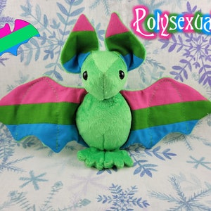 May include: A green plush bat with pink, blue, and green stripes on its wings. The bat is sitting on a white background with a snowflake pattern. The text "Polysexual" is written in pink and white letters above the bat.
