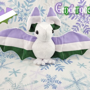 May include: A white plush bat with lavender, white, and green wings. The wings are patterned with the genderqueer pride flag. The bat is sitting on a white background with a snowflake pattern.