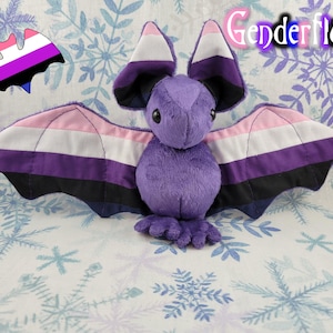 May include: Purple genderfluid bat plush toy with pink, white, purple, and black wings. The wings are made of fabric with the genderfluid pride flag colors.