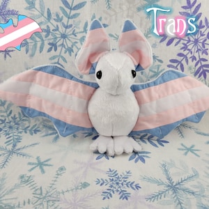 May include: A white plush bat with blue and pink wings featuring the transgender pride flag colors. The bat is sitting on a white background with a blue snowflake pattern. The word "Trans" is written in cursive in the upper right corner of the image.