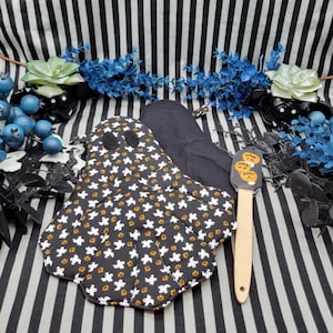 May include: A black and white pot holder with a ghost and pumpkin pattern. The pot holder is shaped like a ghost and has a scalloped edge. A wooden spatula with a pumpkin design is also included.