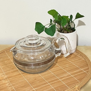 May include: A clear glass teapot with a lid and handle, featuring a brown band around the middle. The teapot sits on a bamboo mat, with a small white potted plant with green leaves in the background.