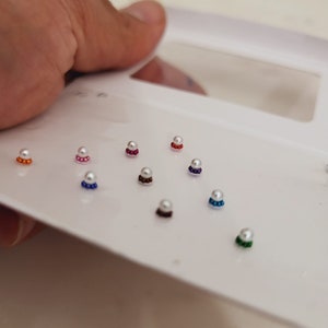 May include: A set of ten white pearl bindi stickers with colored borders. The bindi stickers are arranged in a white plastic case.
