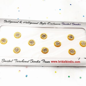 Op de afbeelding: Een verpakking met zeven gouden en witte, met juwelen versierde voorhoofd bindis. De bindis zijn in een rij op een witte achtergrond gerangschikt. De verpakking heeft de tekst "Bollywood & Hollywood Style Exclusive Bridal Bindis" en "Bridal Forehead Bindis From www.bridalbindis.com".