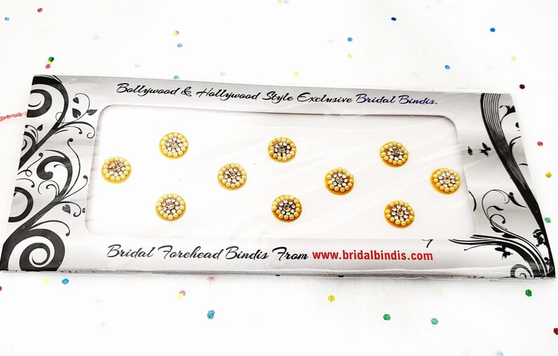 Op de afbeelding: Een verpakking met zeven gouden en witte, met juwelen versierde voorhoofd bindis. De bindis zijn in een rij op een witte achtergrond gerangschikt. De verpakking heeft de tekst "Bollywood & Hollywood Style Exclusive Bridal Bindis" en "Bridal Forehead Bindis From www.bridalbindis.com".