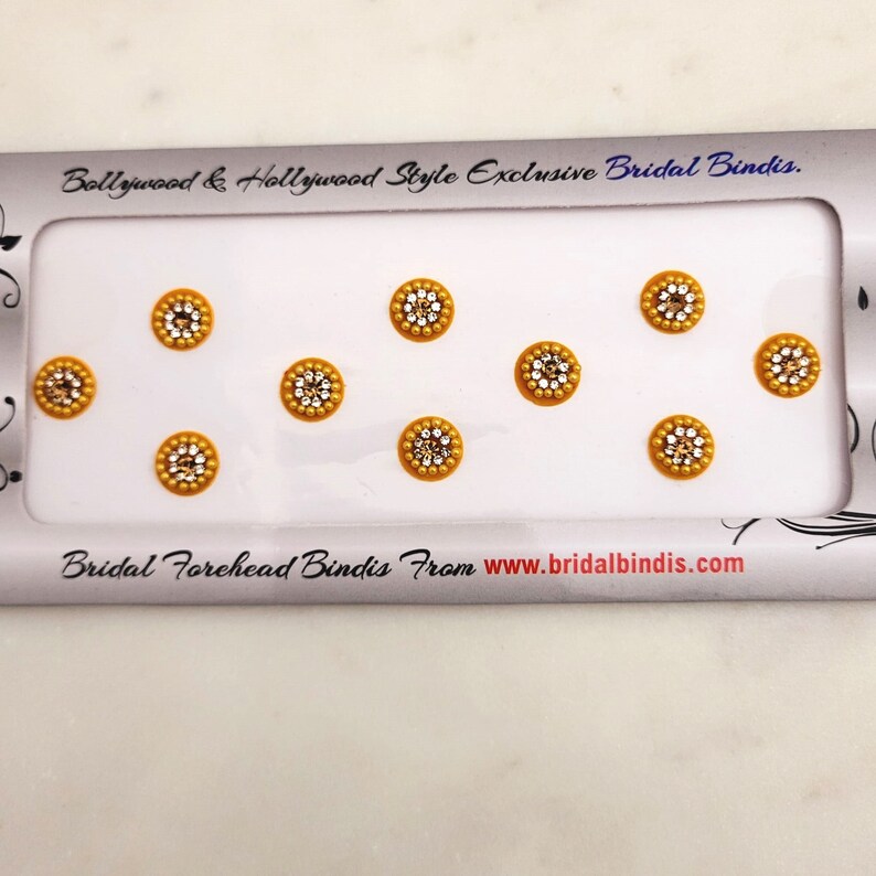 Op de afbeelding: Een vel met acht gouden en transparante strass-bindi-stickers. De bindi-stickers zijn gerangschikt in twee rijen van vier. Het vel is gelabeld met "Bollywood & Hollywood Style Exclusive Bridal Bindis." en "Bridal Forehead Bindis From www.bridalbindis.com".