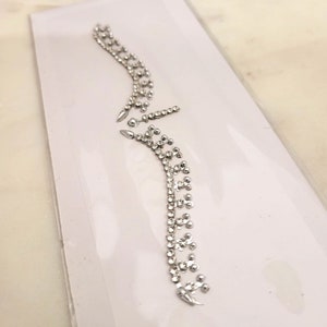 Könnte beinhalten: Eine Folie mit durchsichtigen, selbstklebenden Strasssteinen in Silber. Die Strasssteine sind in zwei geschwungenen Linien angeordnet, eine über der anderen.