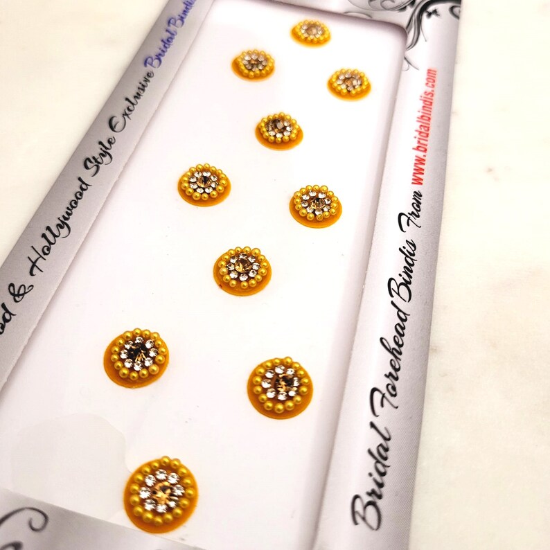 Op de afbeelding: Een vel met acht gouden en heldere strass-voorhoofd bindis. De bindis zijn gerangschikt in twee rijen van vier. Het vel is gelabeld "Bridal Forehead Bindis From www.bridalbindis.com".