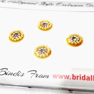 Op de afbeelding: Een vel met acht gele en zilveren zelfklevende bindis. De bindis zijn gerangschikt in twee rijen van vier. De bindis zijn rond en hebben een klein, zilveren, glinsterend midden omgeven door een cirkel van kleine, witte parels.