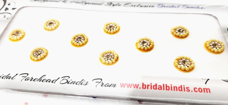 Op de afbeelding: Een vel met acht gele en zilveren zelfklevende bindis. De bindis zijn gerangschikt in twee rijen van vier. De bindis zijn rond en hebben een klein, zilveren, glinsterend midden omgeven door een cirkel van kleine, witte parels.