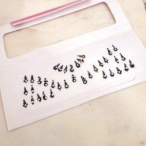 May include: A sheet of white paper with black and white bindi stickers. The stickers feature various designs with small colourful gems.