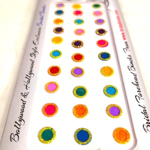 May include: A sheet of colourful, round bindi forehead stickers. The stickers are in various colours, including pink, blue, green, orange, and purple, with gold glitter edging. The packaging reads "Bollywood & Hollywood Style Exclusive Bridal Bindis."