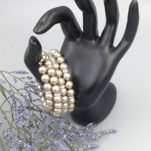 May include: A multi-strand pearl bracelet with small clear rhinestones. The bracelet is made of ivory-coloured pearls and is displayed on a black hand.