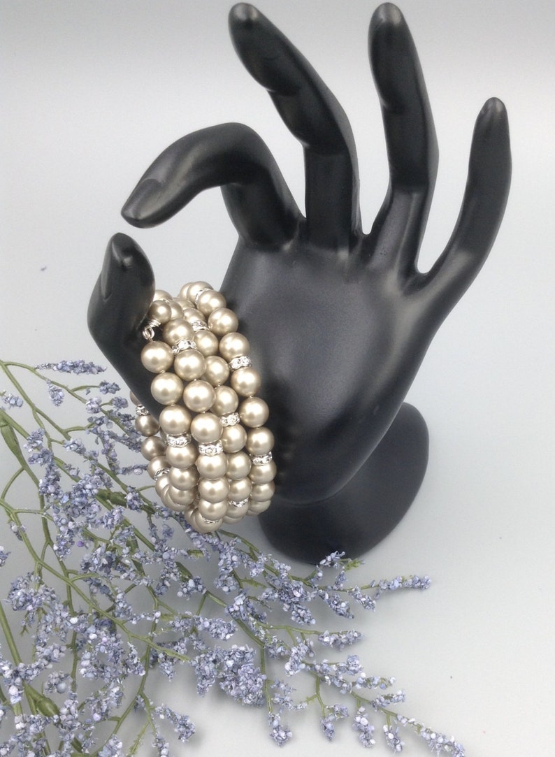 May include: A multi-strand pearl bracelet with small clear rhinestones. The bracelet is made of ivory-coloured pearls and is displayed on a black hand.