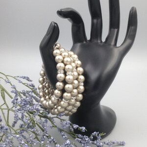 May include: A multi-strand pearl bracelet with a row of clear rhinestones between each strand of pearls. The bracelet is displayed on a black hand-shaped stand.