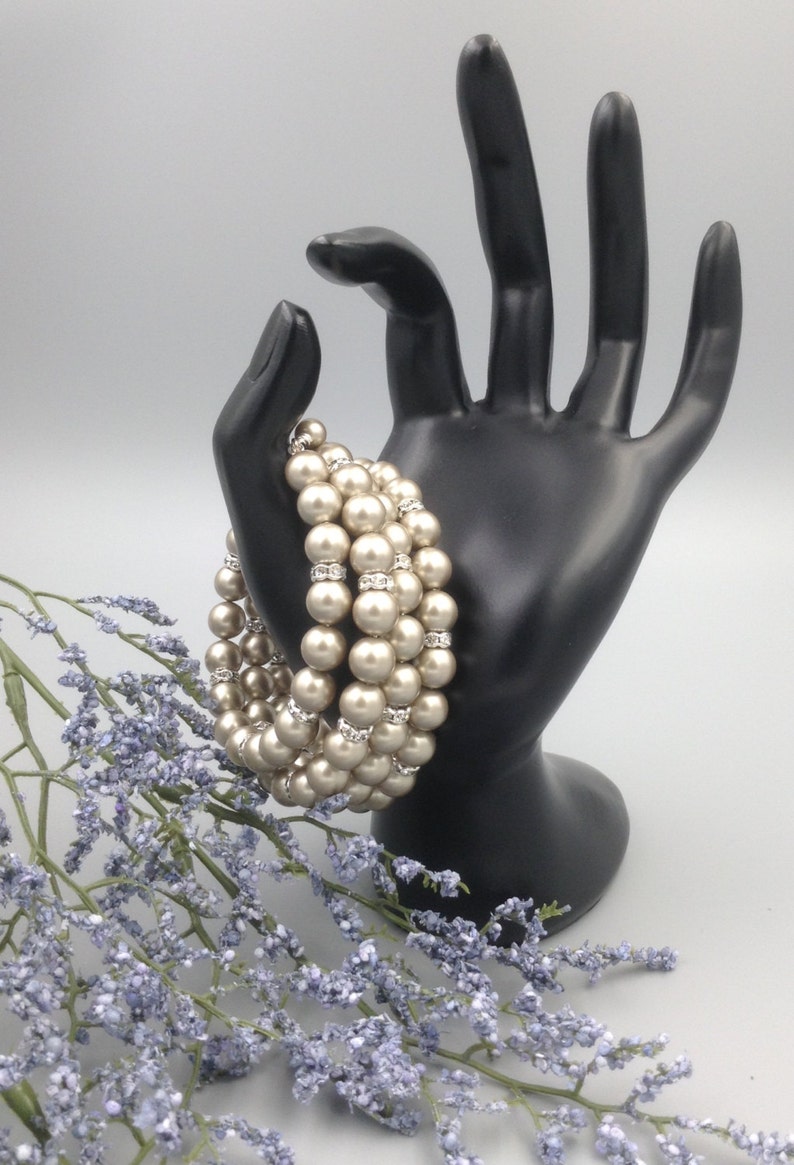 May include: A multi-strand pearl bracelet with a row of clear rhinestones between each strand of pearls. The bracelet is displayed on a black hand-shaped stand.