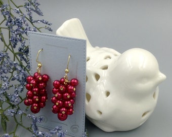 May include: A pair of red beaded earrings with gold hooks. The earrings are shaped like a cluster of grapes and are displayed on a white card.