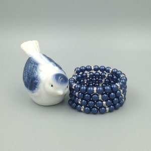 May include: A blue and white porcelain bird figurine next to a multi-strand bracelet made of dark blue faux pearls with clear rhinestone accents.