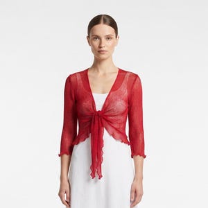 May include: A bright red, sheer, three-quarter sleeve bolero cardigan with a ruffled front tie closure, worn over a simple, sleeveless, knee-length white dress. The outfit is completed with a pair of plain white trainers.
