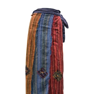 Op de afbeelding: Een kleurrijke wikkelrok met verticale strepen in oranje, blauw en rood. De rok heeft ruitvormige patches met geborduurde details en een gefranjerde zoom. De tailleband is van donkerblauwe stof.