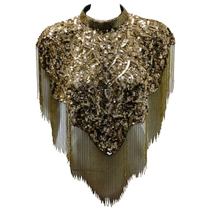 May include: A gold sequined top with a high collar and fringe detailing. The top is heavily embellished with sequins and beads, creating a shimmering effect. The fringe hangs from the shoulders and the bottom hem, adding movement.