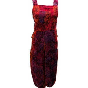 Op de afbeelding: Een levendige jumpsuit met bloemenpatroon in tinten rood, paars en zwart. De jumpsuit heeft een vierkante halslijn, brede bandjes en een geplooide taille. De stof heeft een fluweelzachte textuur en het algehele ontwerp is bohemien.