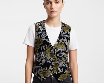 Op de afbeelding: Een zwart vest met een bloemenpatroon in olijfgroene en zilveren tinten. Het kledingstuk heeft een V-hals, een knoopsluiting aan de voorzijde en twee kleine zakken. Het wordt gedragen over een effen wit T-shirt met korte mouwen.