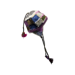 Pode incluir: Um chapéu de malha patchwork colorido com protetores de orelhas e longas amarrações trançadas que terminam em pequenos pompons. O chapéu apresenta quadrados cinza, azul, roxo, verde e rosa. As amarrações são verde escuro e os pompons são rosa.