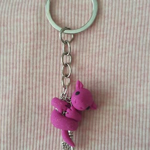 May include: A pink keychain featuring a small, glittery, stylized animal figure. The figure is attached to a silver chain and keyring. The background is a light pink fabric with a ribbed pattern.