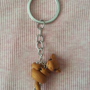 May include: A handmade keychain featuring a small, brown, sculpted animal charm. The charm is attached to a silver-colored chain and keyring. The background is a pink and white striped fabric.