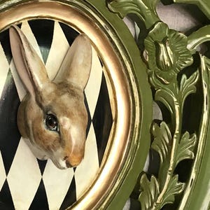 May include: A decorative wall hanging featuring a rabbit head emerging from a circular frame with a black and white diamond pattern. The frame is gold-trimmed and set within a larger, ornate green frame with floral accents.