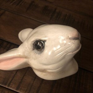 May include: White ceramic rabbit figurine with a pink nose and black eyes. The rabbit's head is tilted to the side, and its ears are perked up.