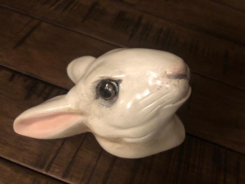 May include: White ceramic rabbit figurine with a pink nose and black eyes. The rabbit's head is tilted to the side, and its ears are perked up.
