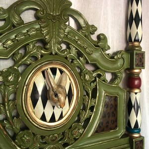 May include: A green, ornate headboard with a rabbit sculpture in a circular frame with a black and white diamond pattern. The headboard has gold accents and a leopard print panel. The bedpost is black and white with a diamond pattern.