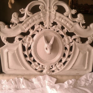 May include: A white, ornate headboard with carved floral and scroll details. A circular inset features a white rabbit head. The headboard is positioned above white ruffled bedding, creating a vintage aesthetic.