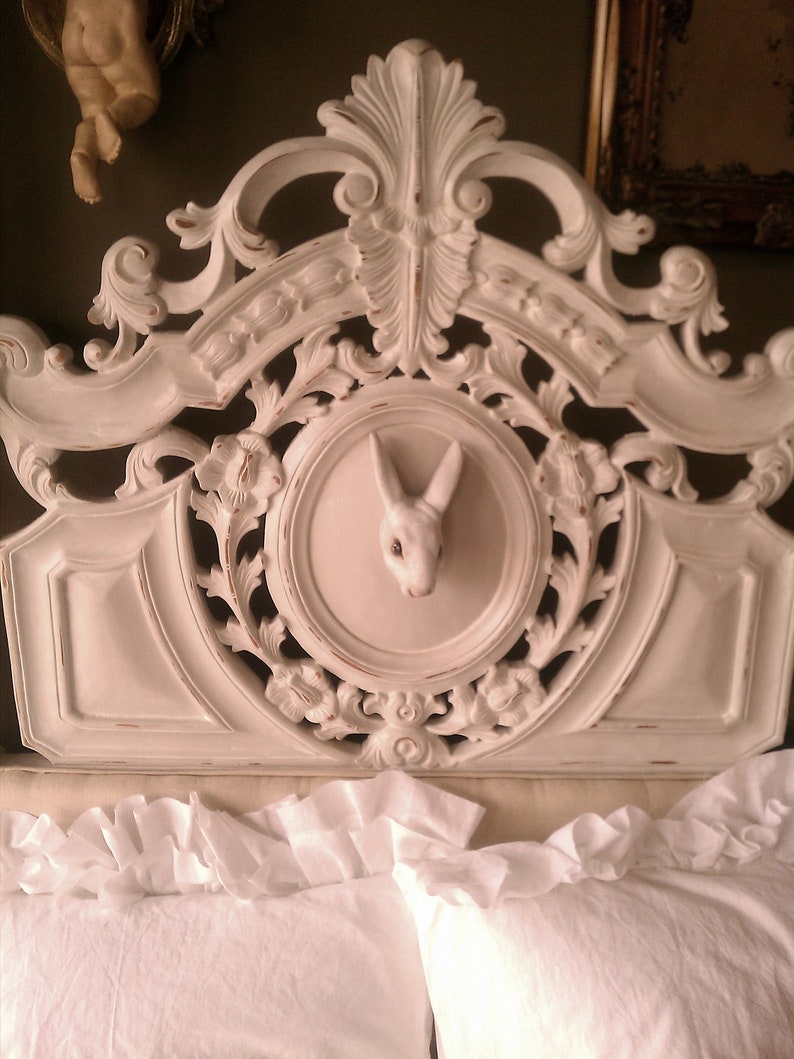 May include: A white, ornate headboard with carved floral and scroll details. A circular inset features a white rabbit head. The headboard is positioned above white ruffled bedding, creating a vintage aesthetic.
