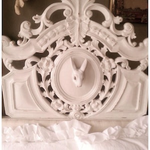May include: White ornate headboard with a carved rabbit head in the center. The headboard is made of wood and has a distressed white finish.