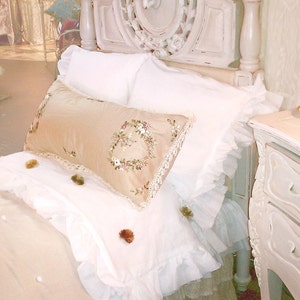 May include: A white, ornate bed frame with a white ruffled bedspread and a beige pillow with floral embroidery. The bed is decorated with a white lace trim and a white ruffled skirt.