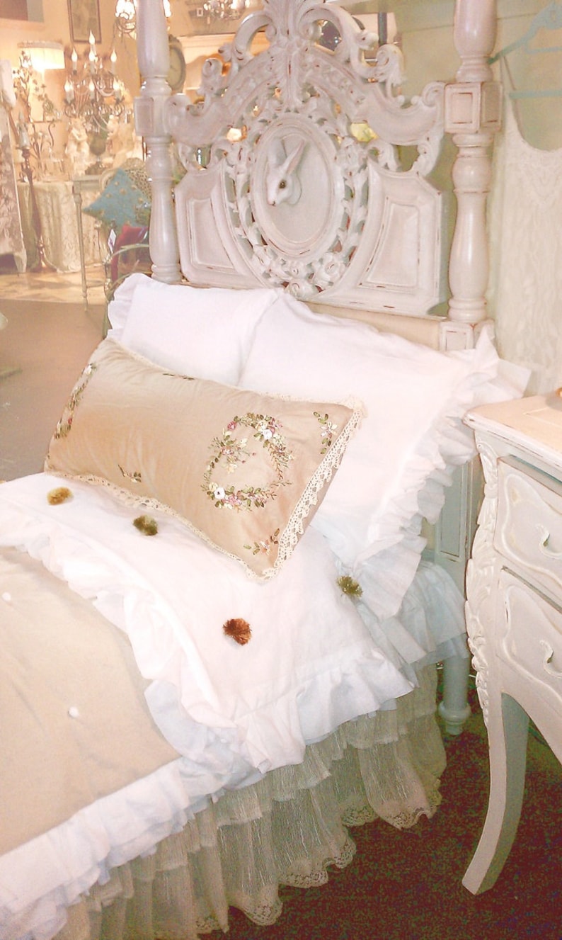 May include: A white, ornate bed frame with a white ruffled bedspread and a beige pillow with floral embroidery. The bed is decorated with a white lace trim and a white ruffled skirt.